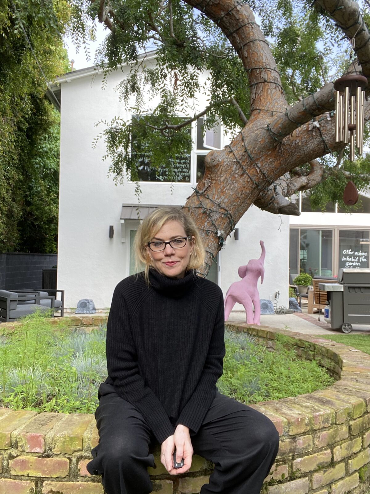 A woman in a black turtleneck sits on a raised brick garden bed