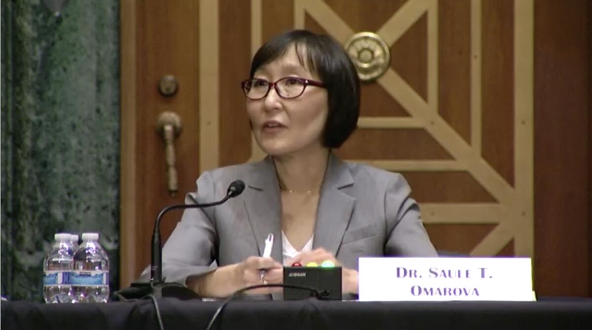Saule T. Omarova sits at a table with a microphone on it and a door behind her