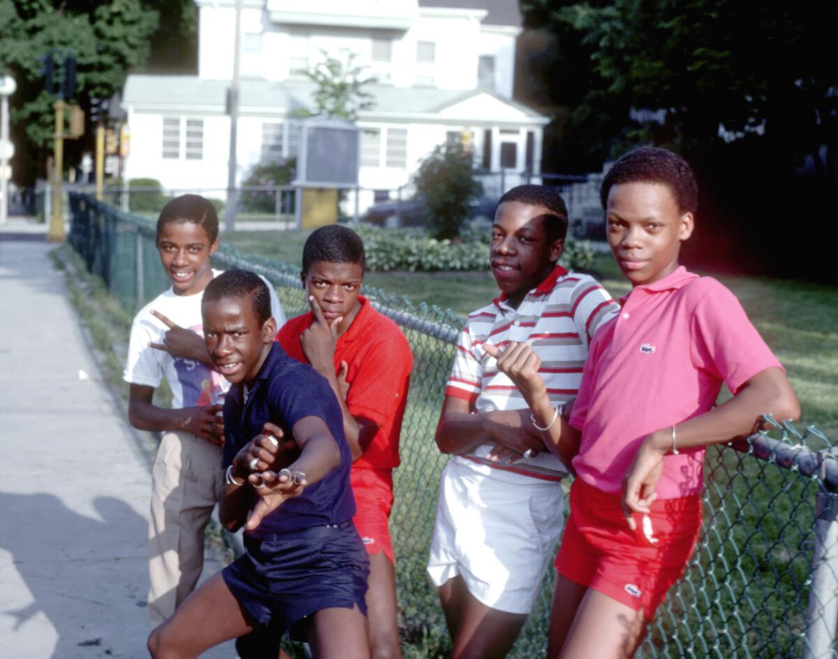 Five young teenagers stand in front of a chain-link fence in the 1980s.