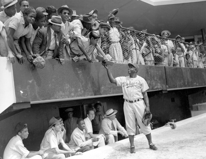 A man in a baseball uniform and holding a glove in one hand hands a notebook to a fan among a crowd in the stands.