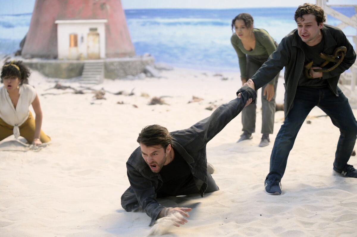 On a beach, two women in the background crouch. Two men in the foreground, one crawling and screaming, one holding him back