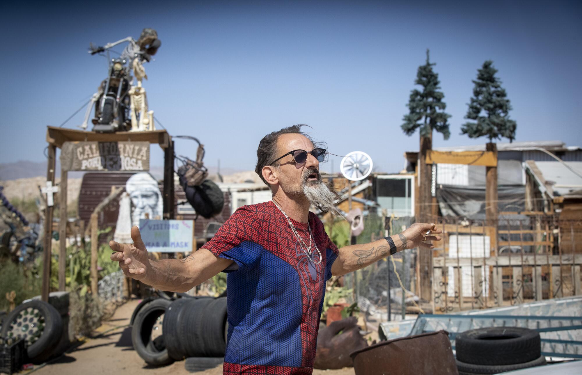 Wearing a red-and-blue Spider-Man T-shirt, Rodney "Spyder" Wild holds his arms out wide at his space in Slab City. 