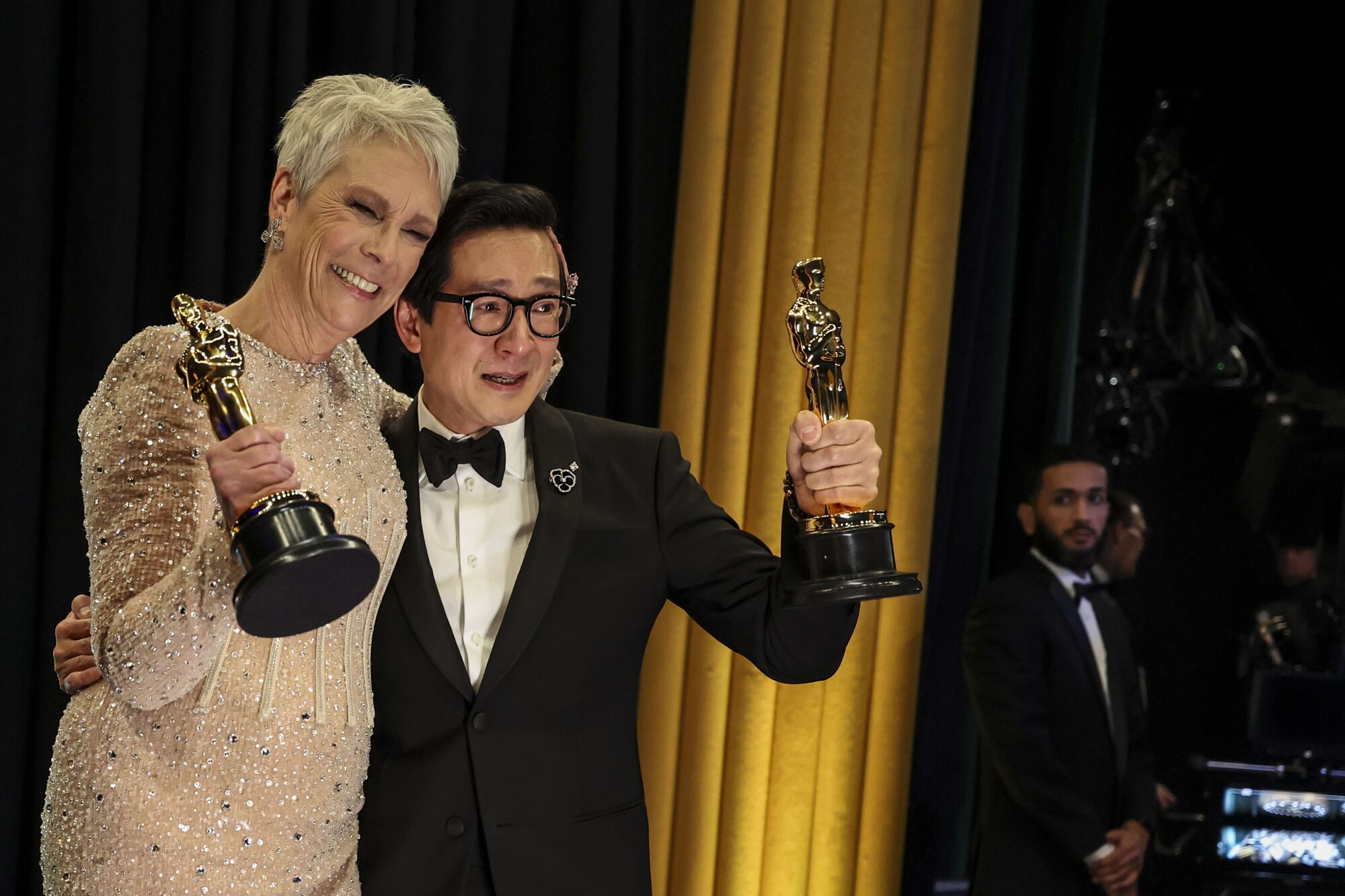Woman in champagne dress and man in tuxedo both hold up Oscars 