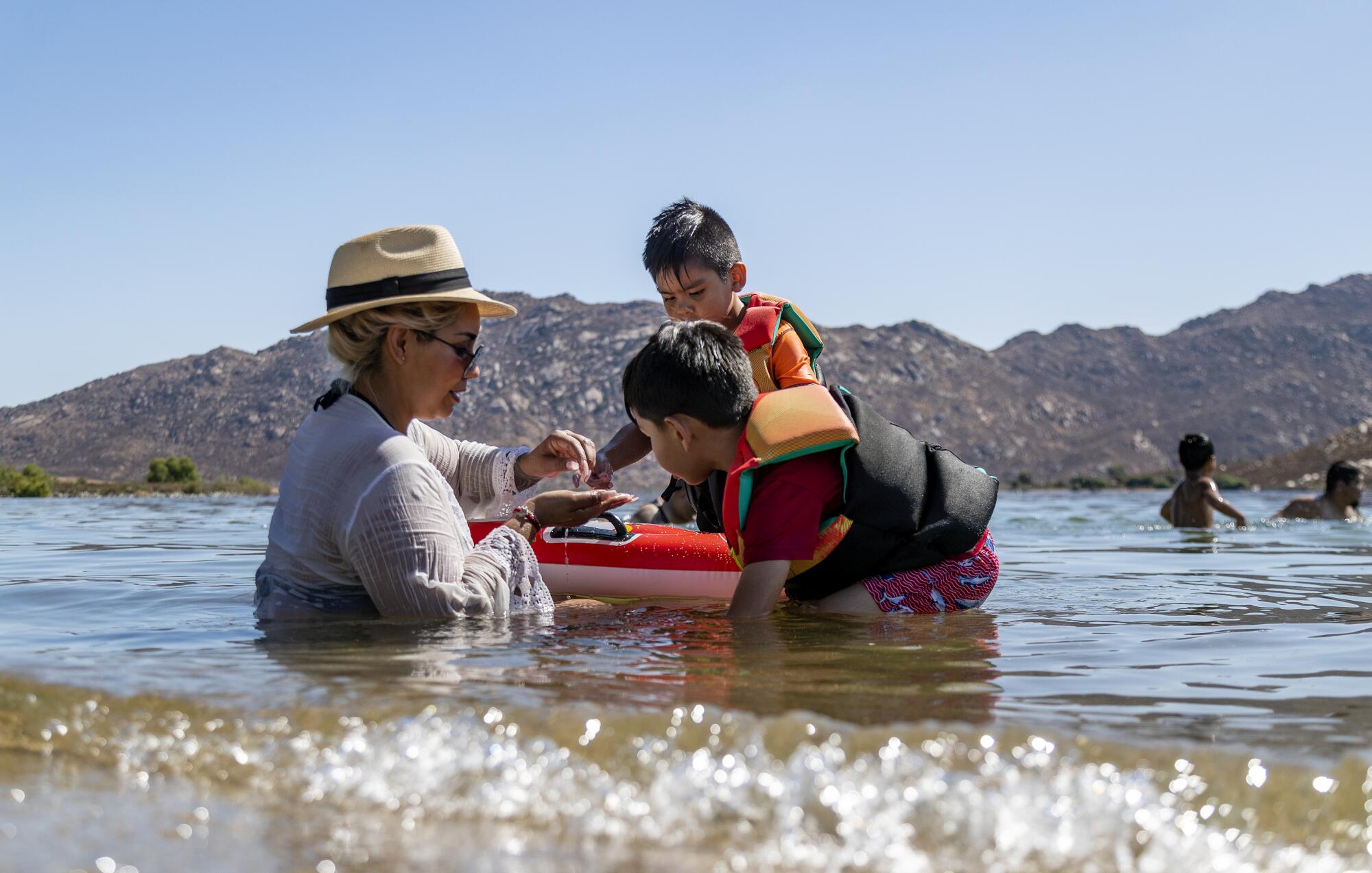 A mom and her young boys in life vests play in the shallow water of a lake