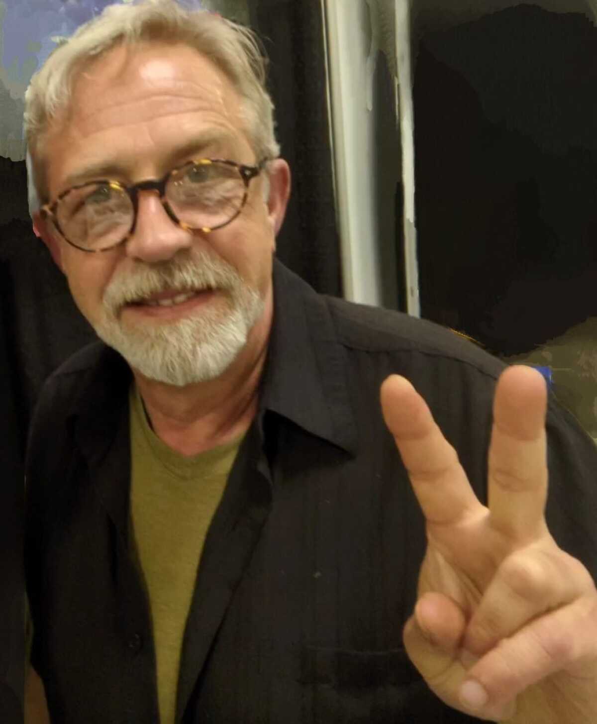A man with gray hair, a beard and glasses making a peace sign.