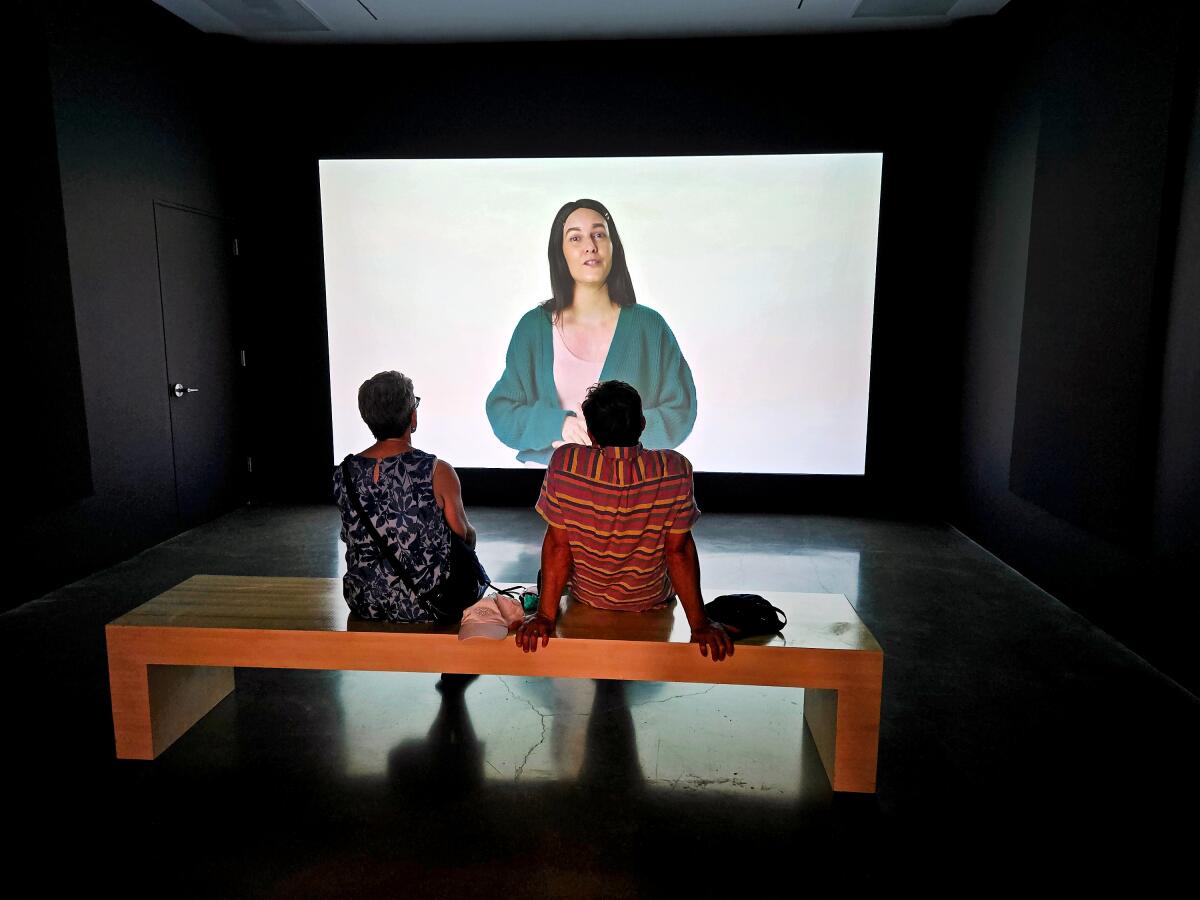 Two people sit on a bench watching a woman projected on the screen before them.