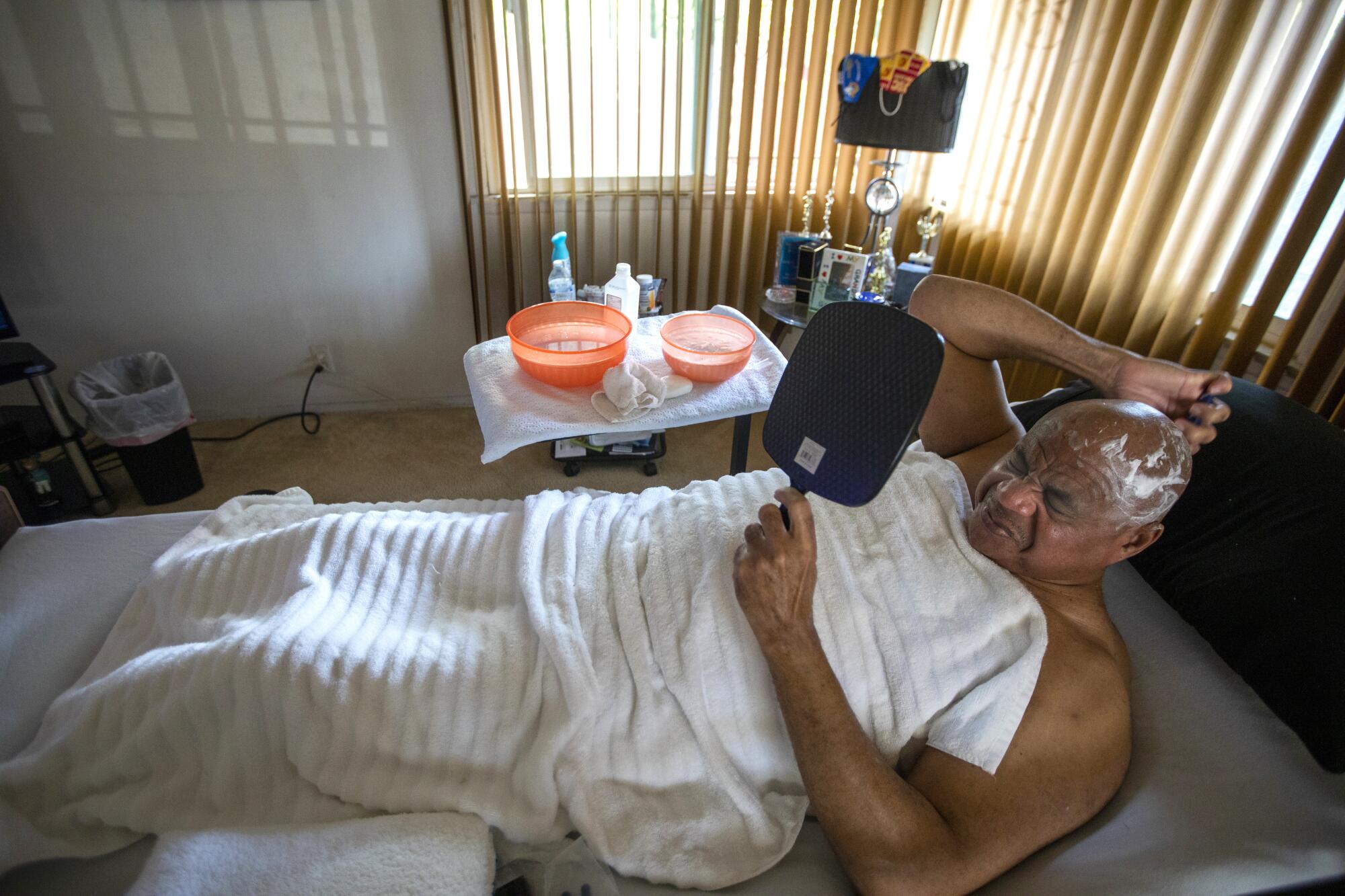 Bill Crawford starts his day, cleaning up and shaving his head in bed in his living room in Watts.