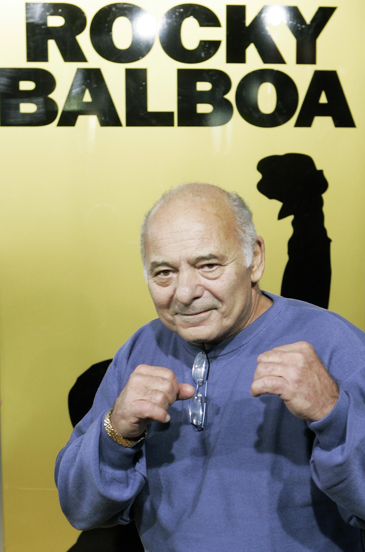Burt Young wears a blue sweatshirt and holds up fighting fists.
