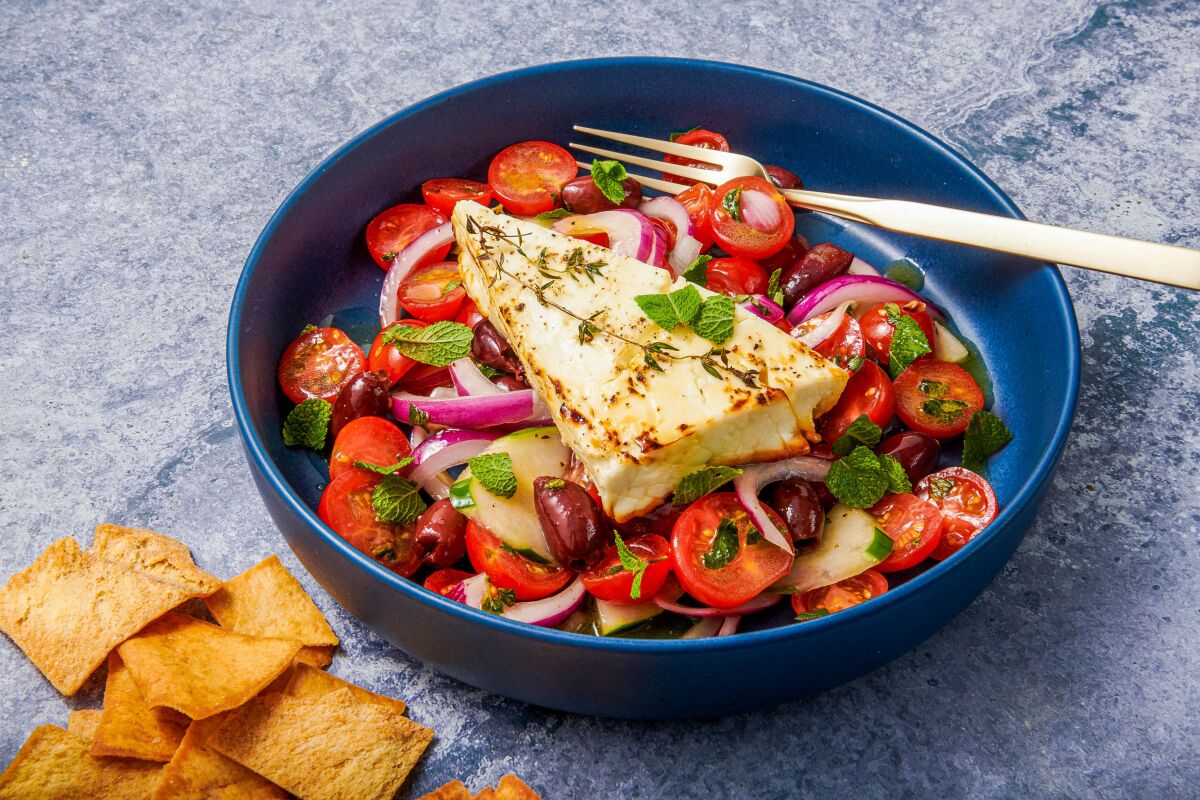A golden slab of grilled feta sits on top of tomatoes, olives and other vegetables.