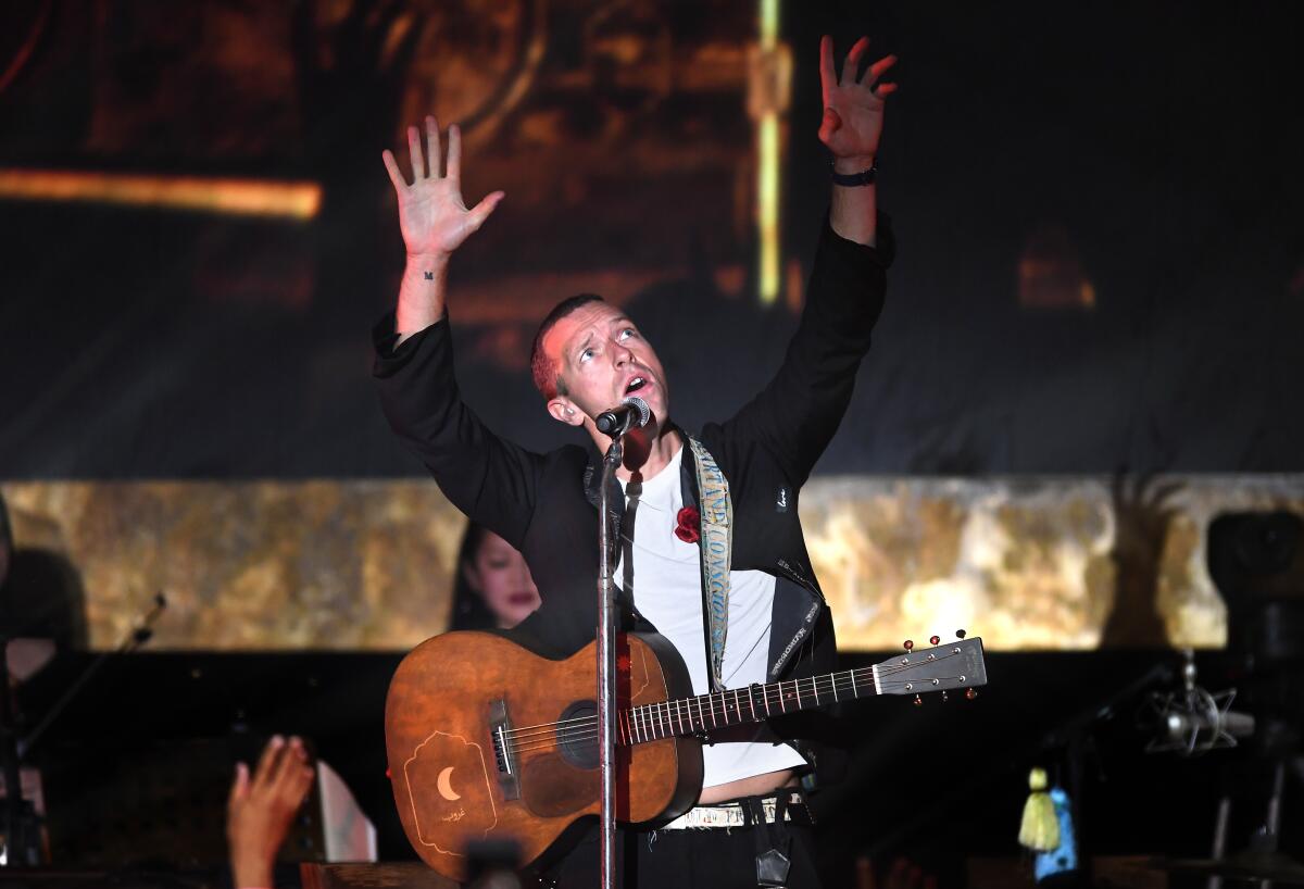 Chris Martin gestures with his arms on a stage with a guitar strapped over his shoulder.