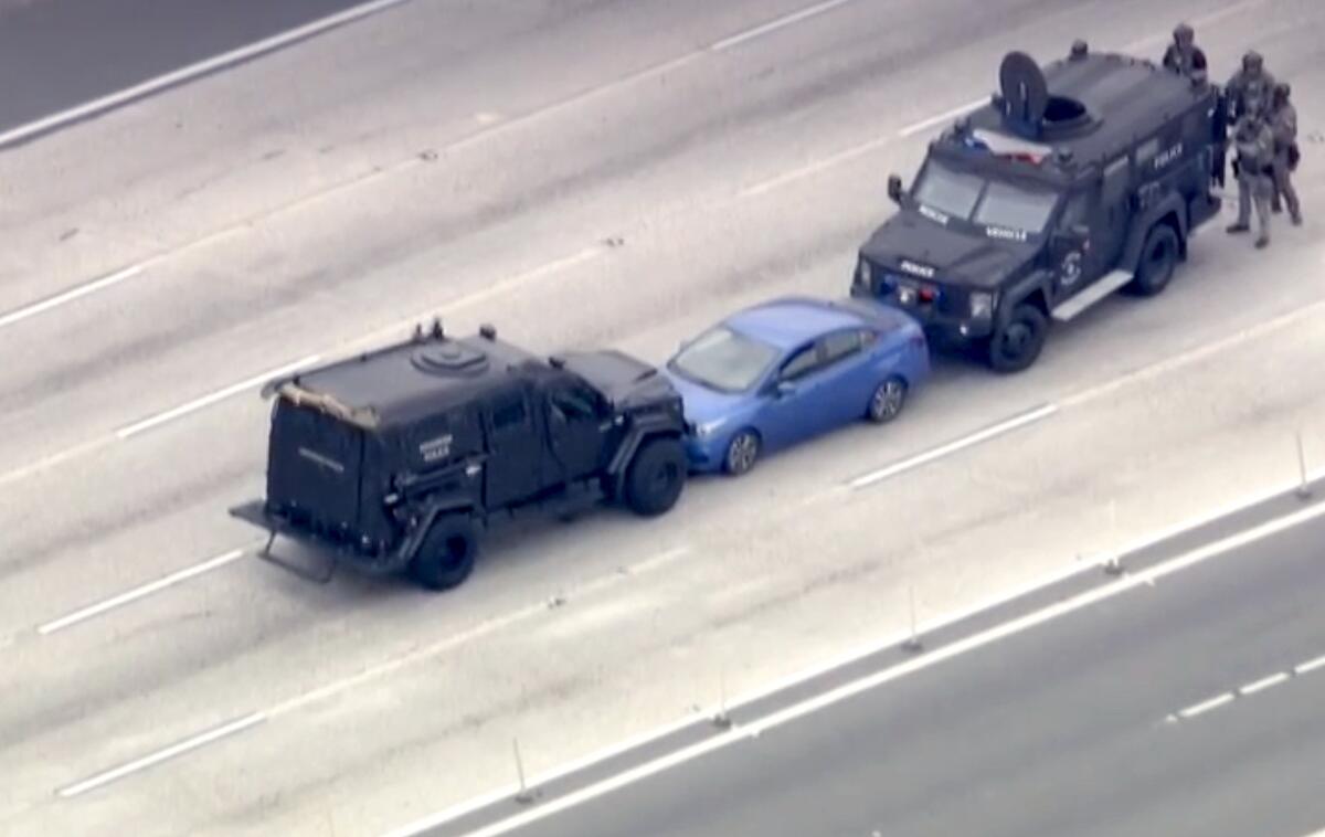 Two large black vehicles box in a blue sedan on the freeway while law enforcement stands behind one black vehicle.