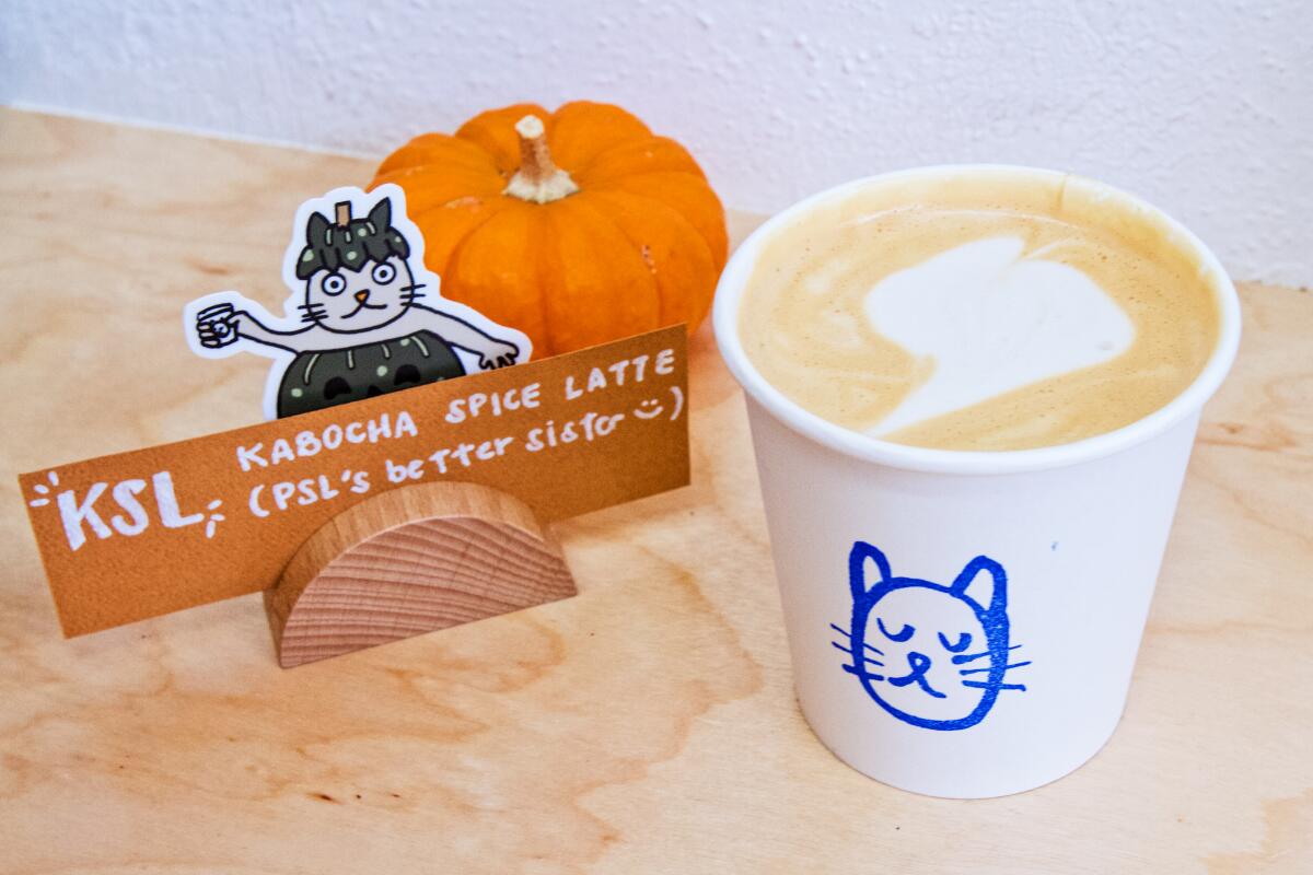 A white paper cup with a blue cat head drawn on it, next to a KSL Kabocha Spice Latte sign with a cat in a pumpkin