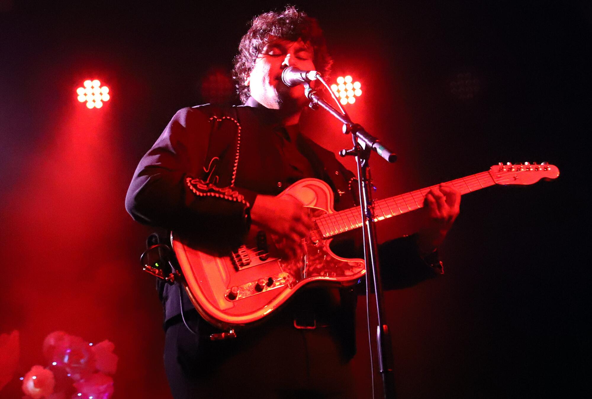 A performer bathed in red lights plays a guitar and sings into a microphone.