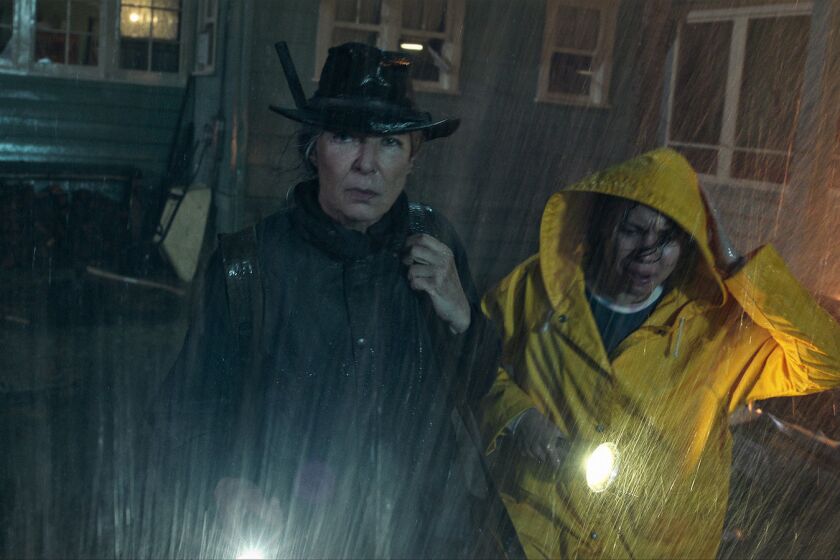 A woman with a rifle and a woman with a flashlight in the rain beside a burning vehicle in the movie "Lou." 