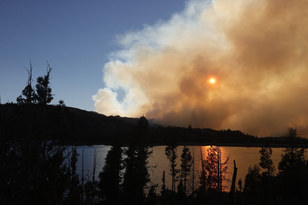 The sun is dimmed by smoke over a lake surrounded by pine trees.