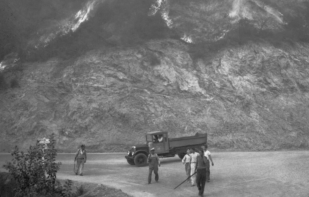 An old-time fire truck sits in front of a burning hillside. Firefighters, mostly in street clothes, walk along a road.