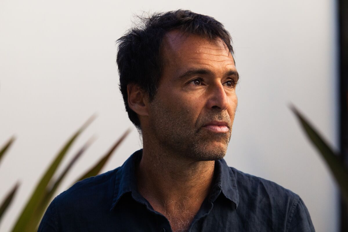 A man in a dark blue button-down shirt stands in front of some fronds.
