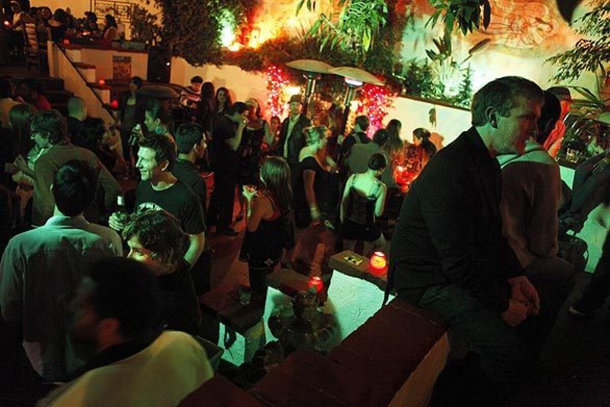 Crowds of people, some holding drinks, chat inside a tropical-themed nightclub.