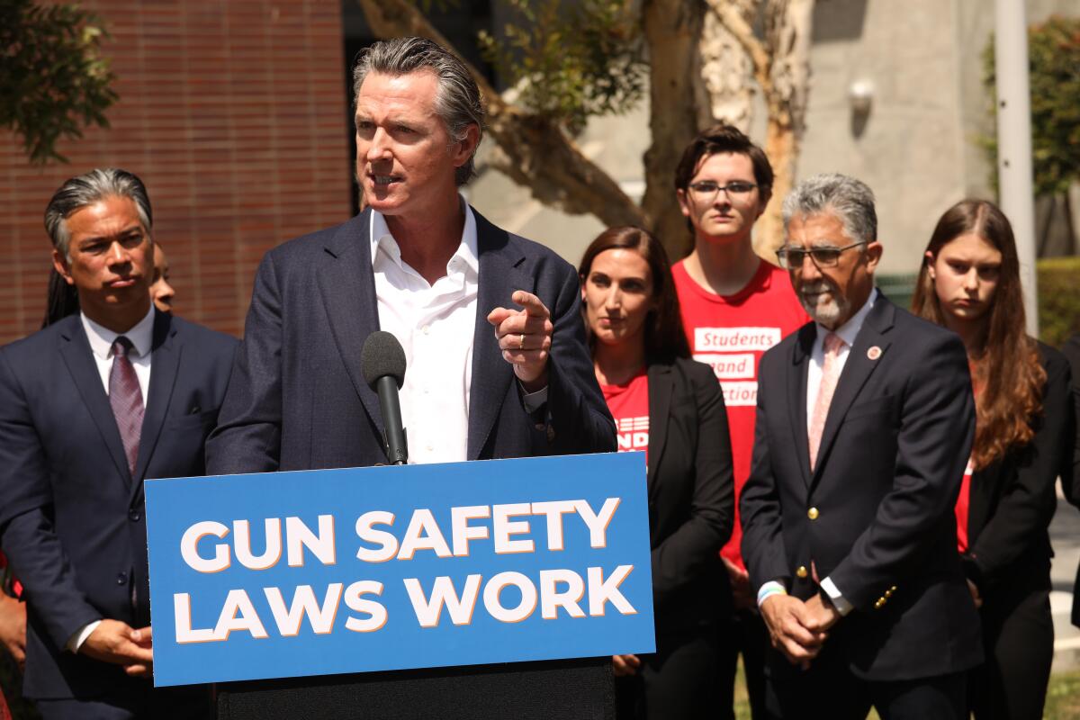 Gavin Newsom points while speaking at a lectern with a sign that says “gun safety laws work” as people stand behind him