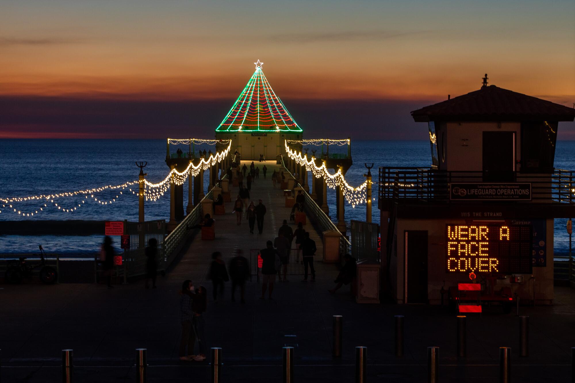 Holiday lights cover a pier at sunset as an electric sign reads "Wear a face cover."