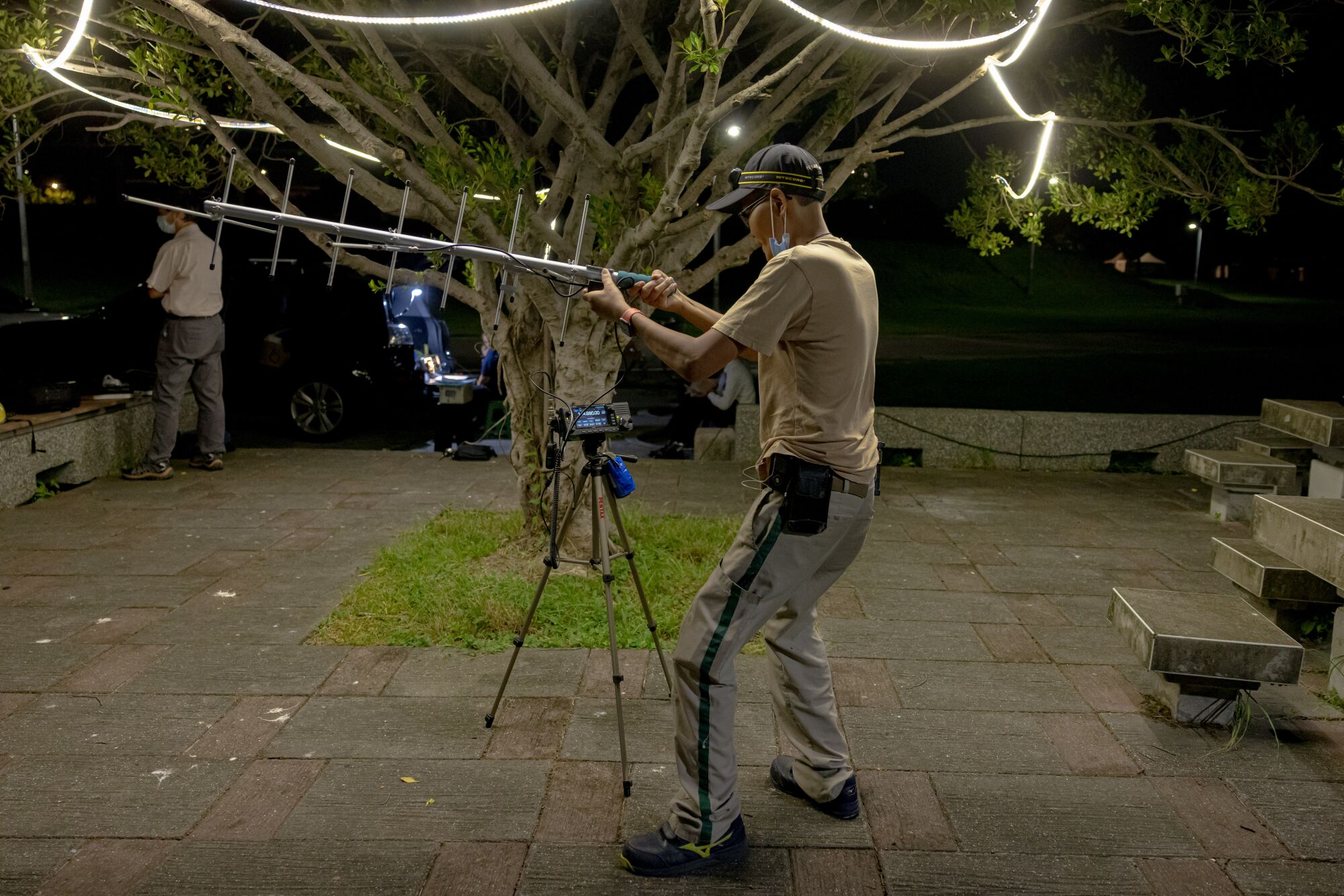 A man in T-shirt, pants and cap assembles an antenna near a tree with lights strung up