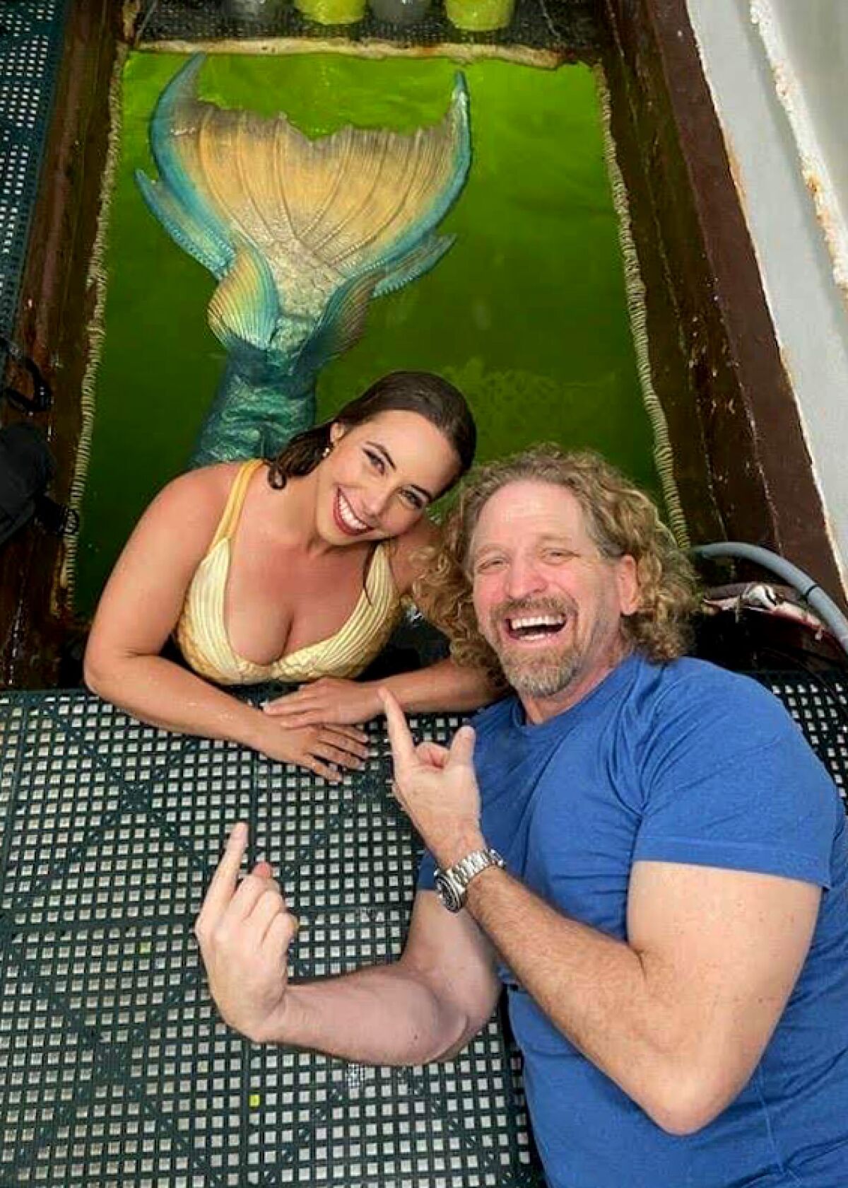 A man pointing to a woman dressed as a mermaid, seen in the green water through a rectangular opening in the floor