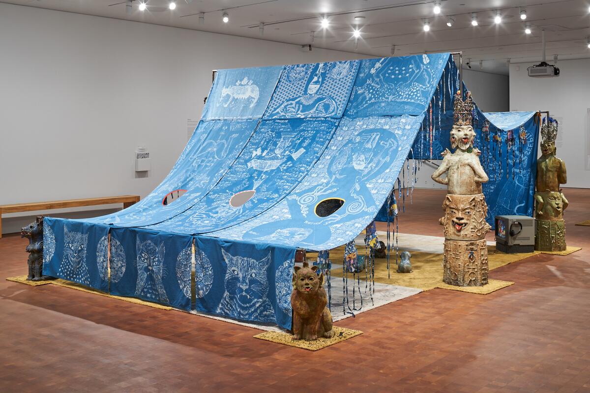 An indigo tent in a museum gallery is surrounded by ceramic cats and strange human figures.