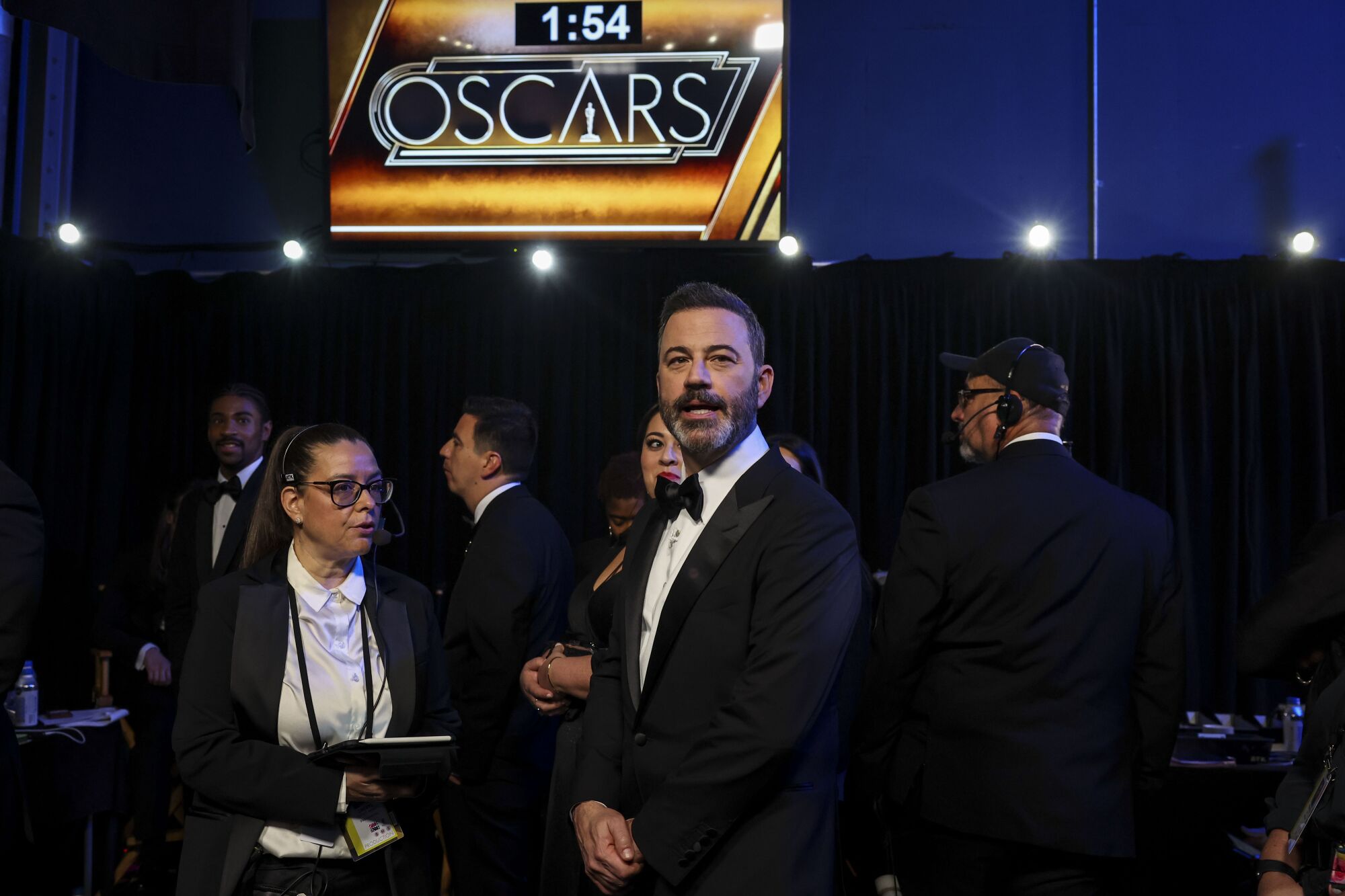 Man in tuxedo under Oscar sign stands among crew members
