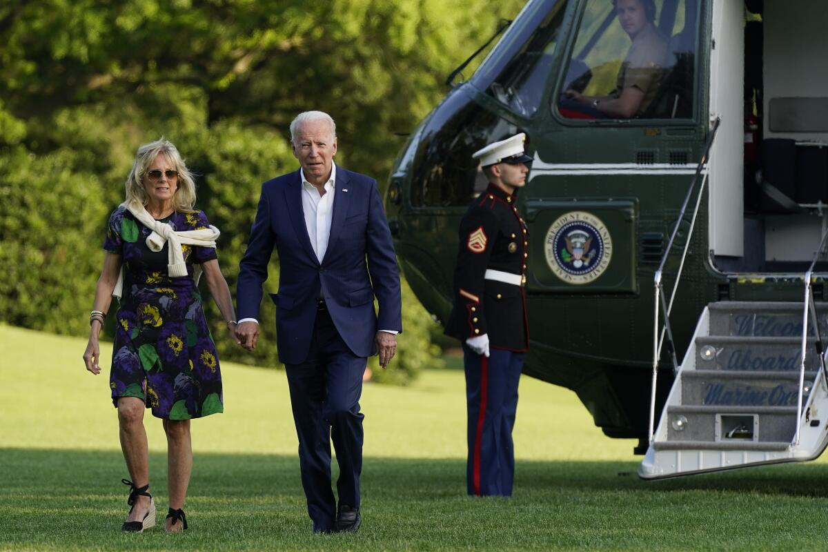 Joe and Jill Biden walk across grass as a man in military uniform stands at attention next to a helicopter.