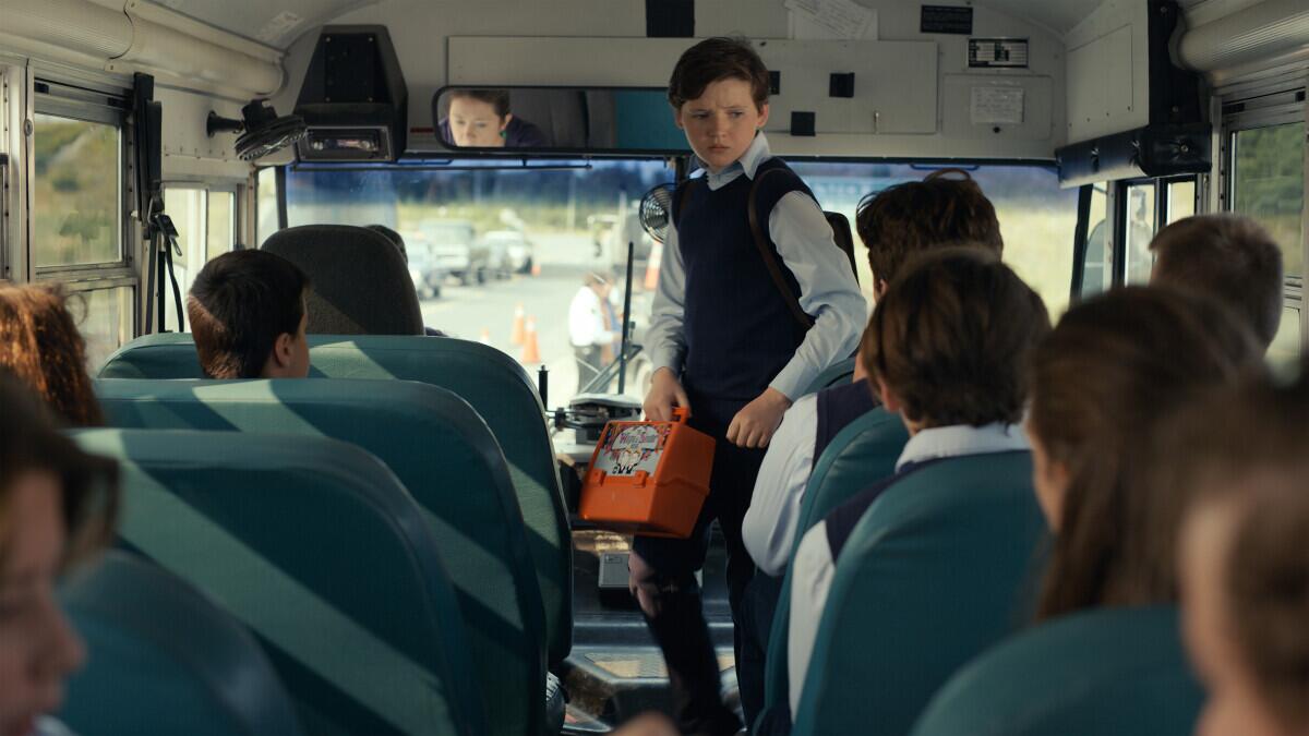 A boy holding an orange lunch box and wearing a long sleeve shirt and sweater vest stands in the aisle of a school bus.
