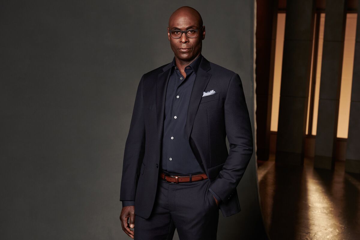 Lance Reddick, who is wearing glasses, stands in front of a gray backdrop in a dark suit and shirt.