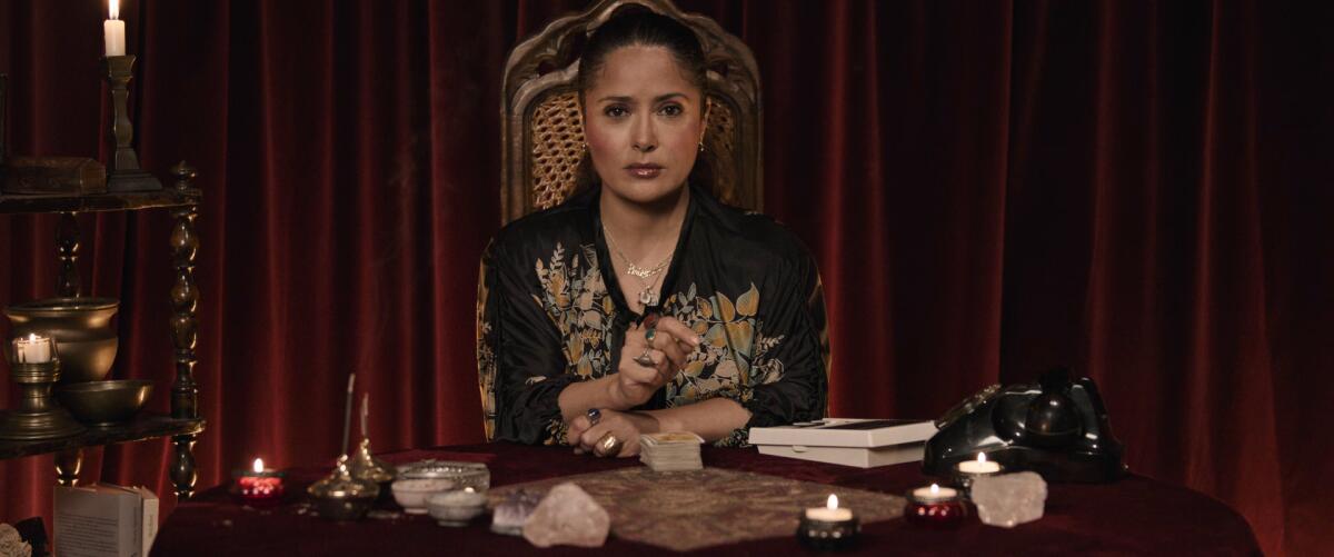 A woman at the head of a candlelit table in front of a red velvet curtain.