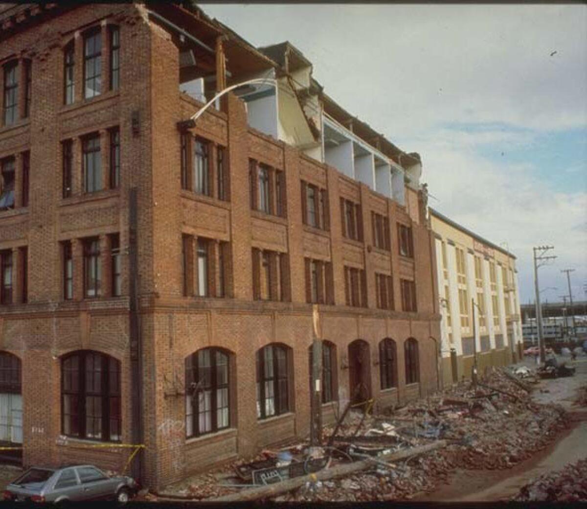 The brick wall at the fourth-floor level of a building is gone. Piles of bricks and other debris fill the sidewalk below.  