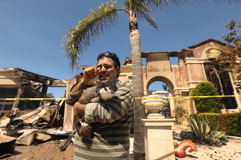 In front of a charred facade, a man wipes an eye as he holds a cat. 