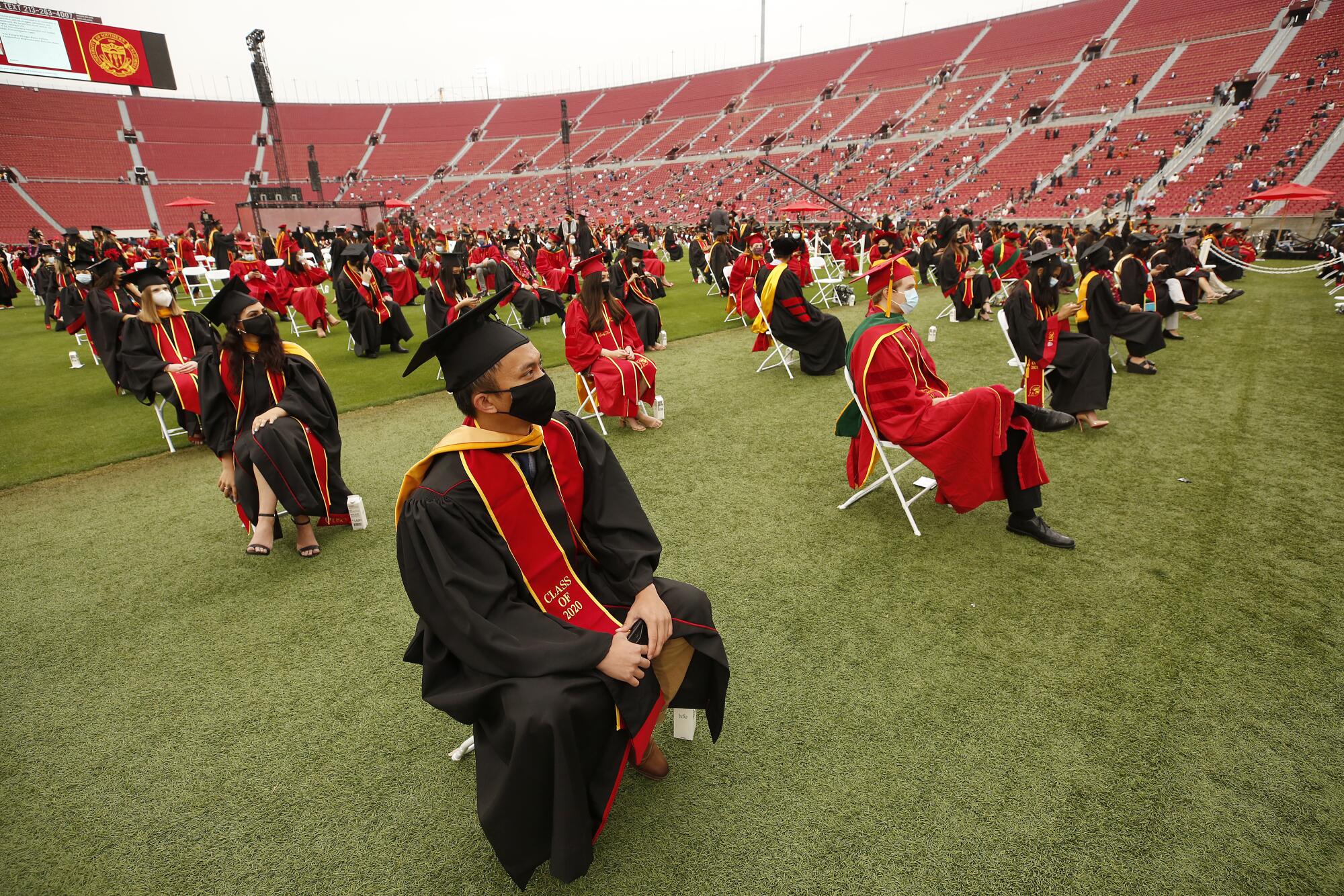 Students in red or black gowns sit on folding chairs spaced out on the USC football field