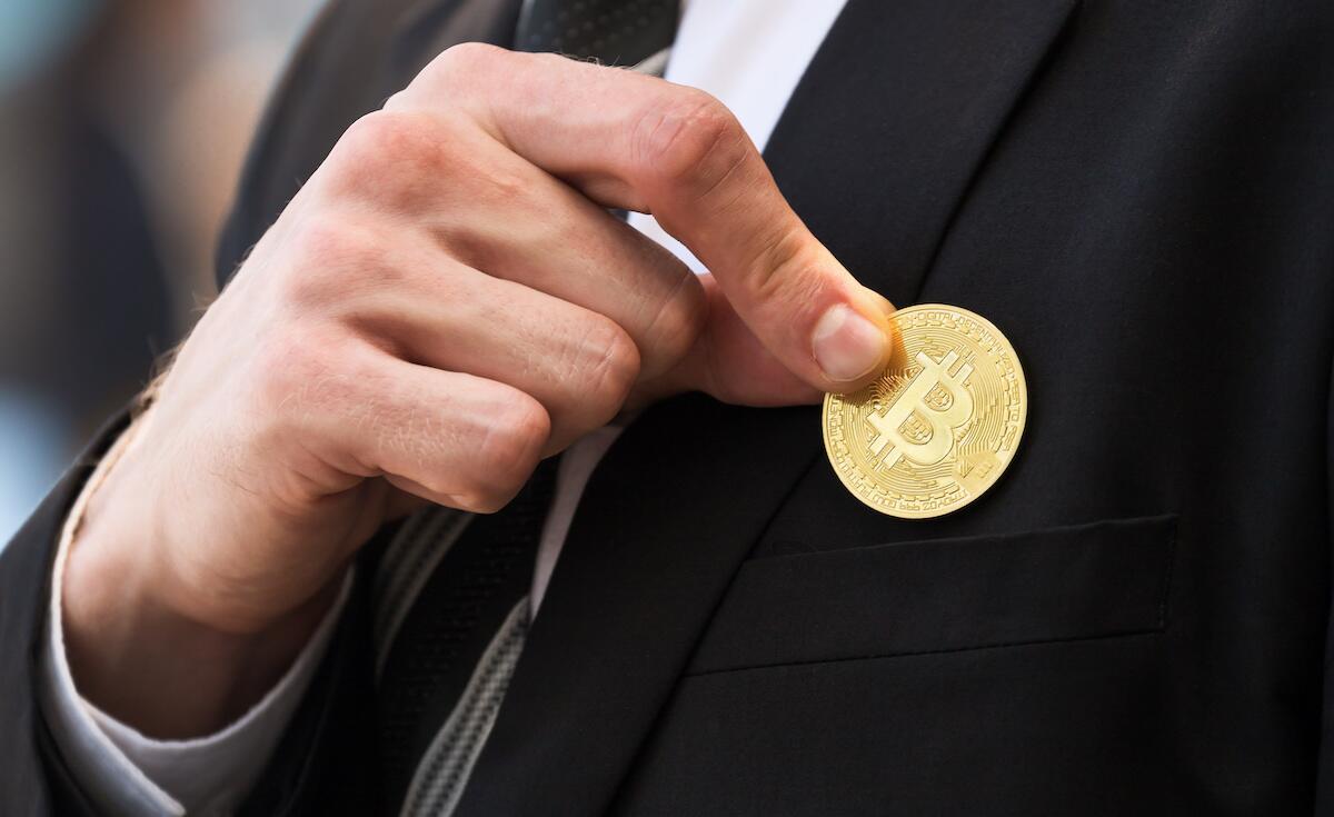 A man in a dark suit holds a gold coin with the Bitcoin logo on it, a B that looks like a dollar sign.