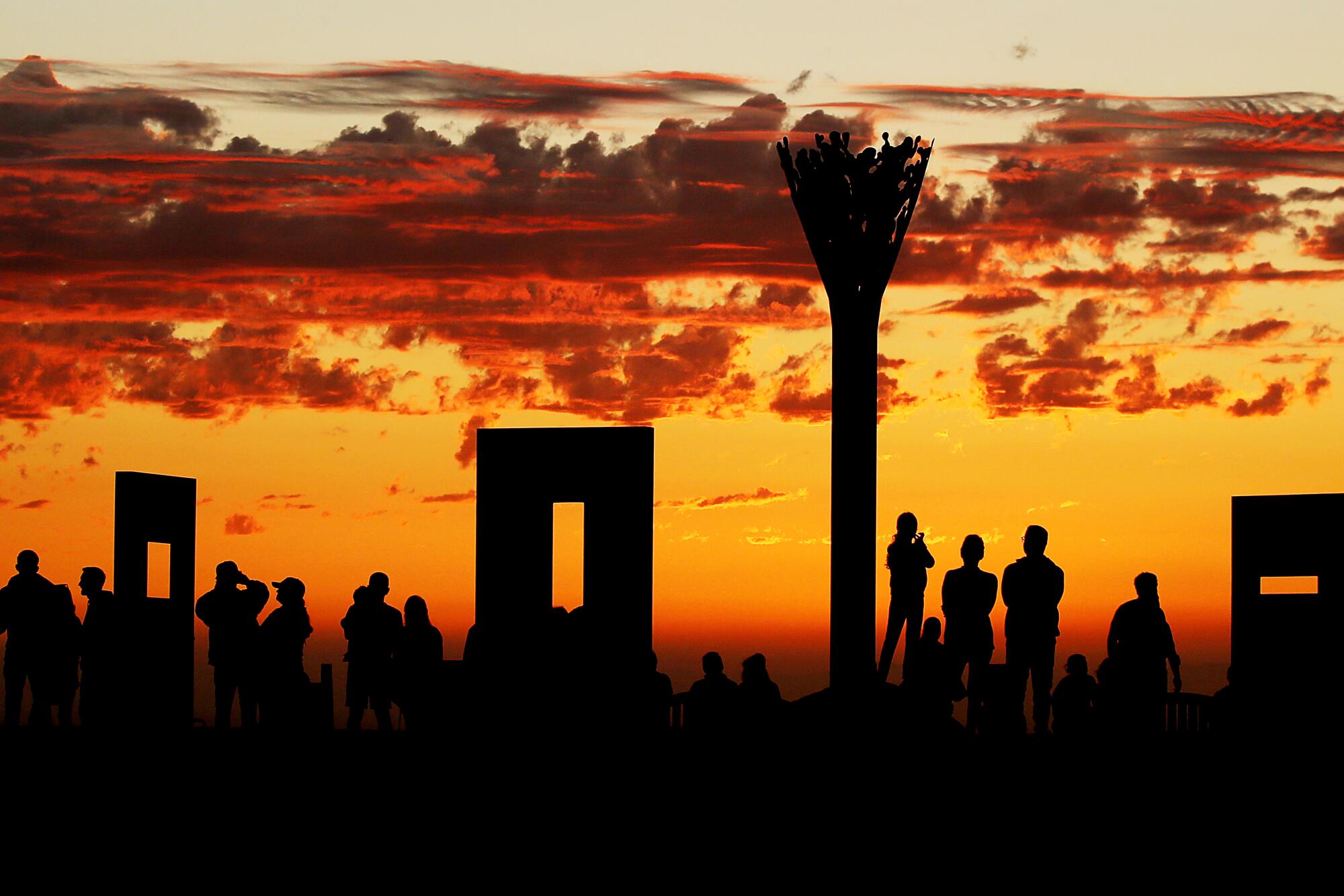 People and objects are silhouetted against an orange sky with clouds.