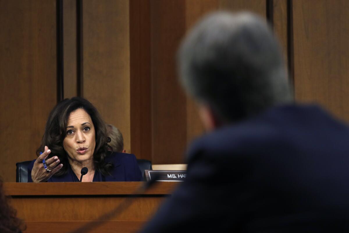 Kamala Harris speaks from the front of a wood-paneled room to Brett Kavanaugh, who's pictured from behind in the foreground