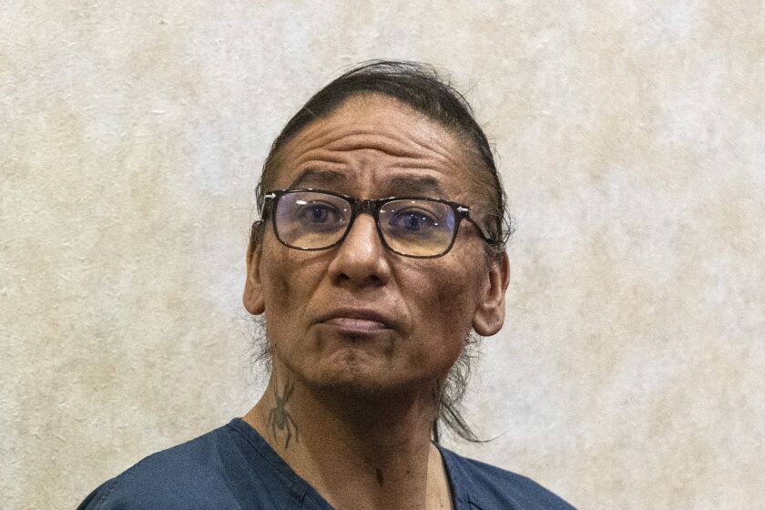 A man with a blue jailhouse shirt, glasses and tattoos on his chin and neck sits in a courtroom