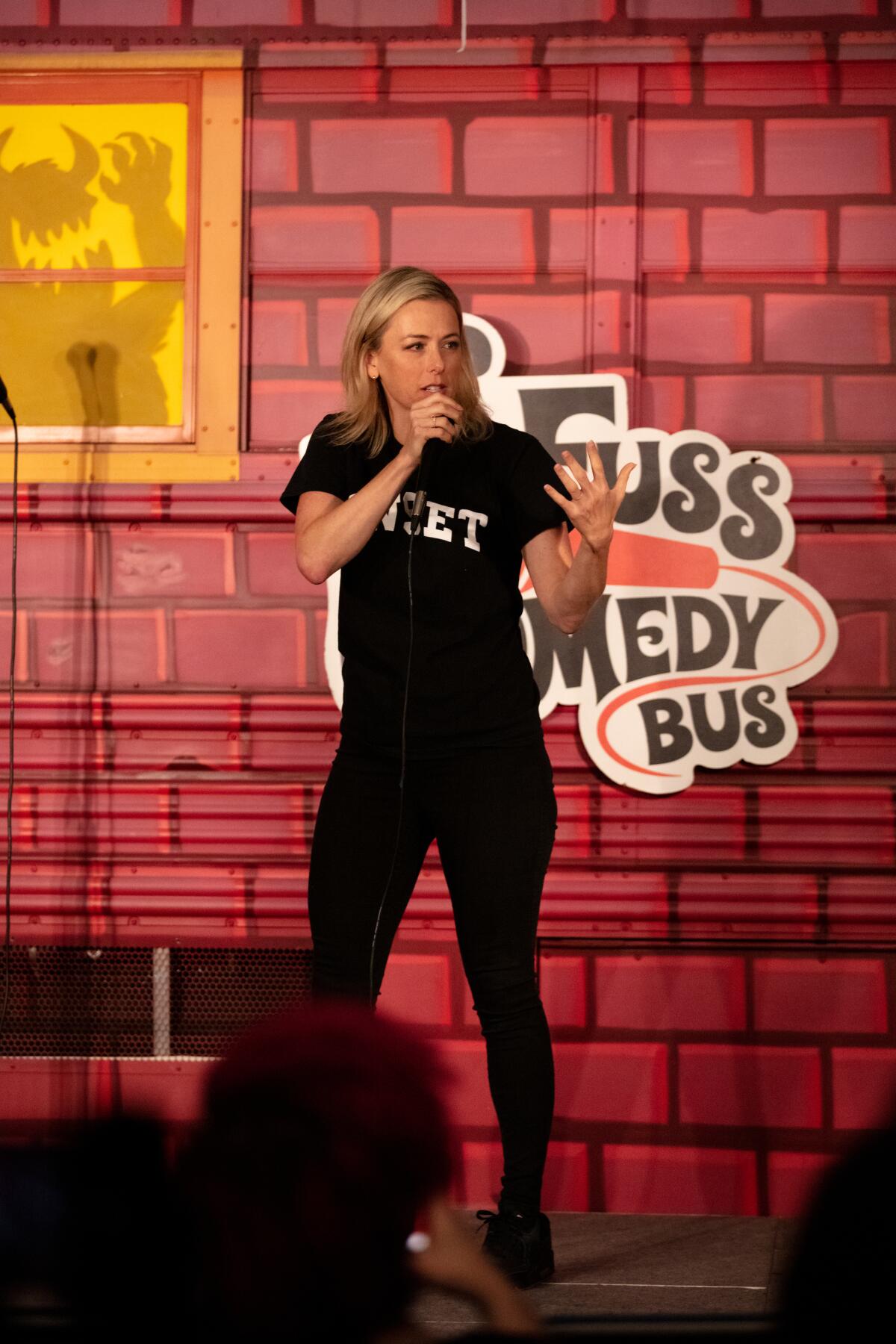 A blond woman wearing all black performs comedy.