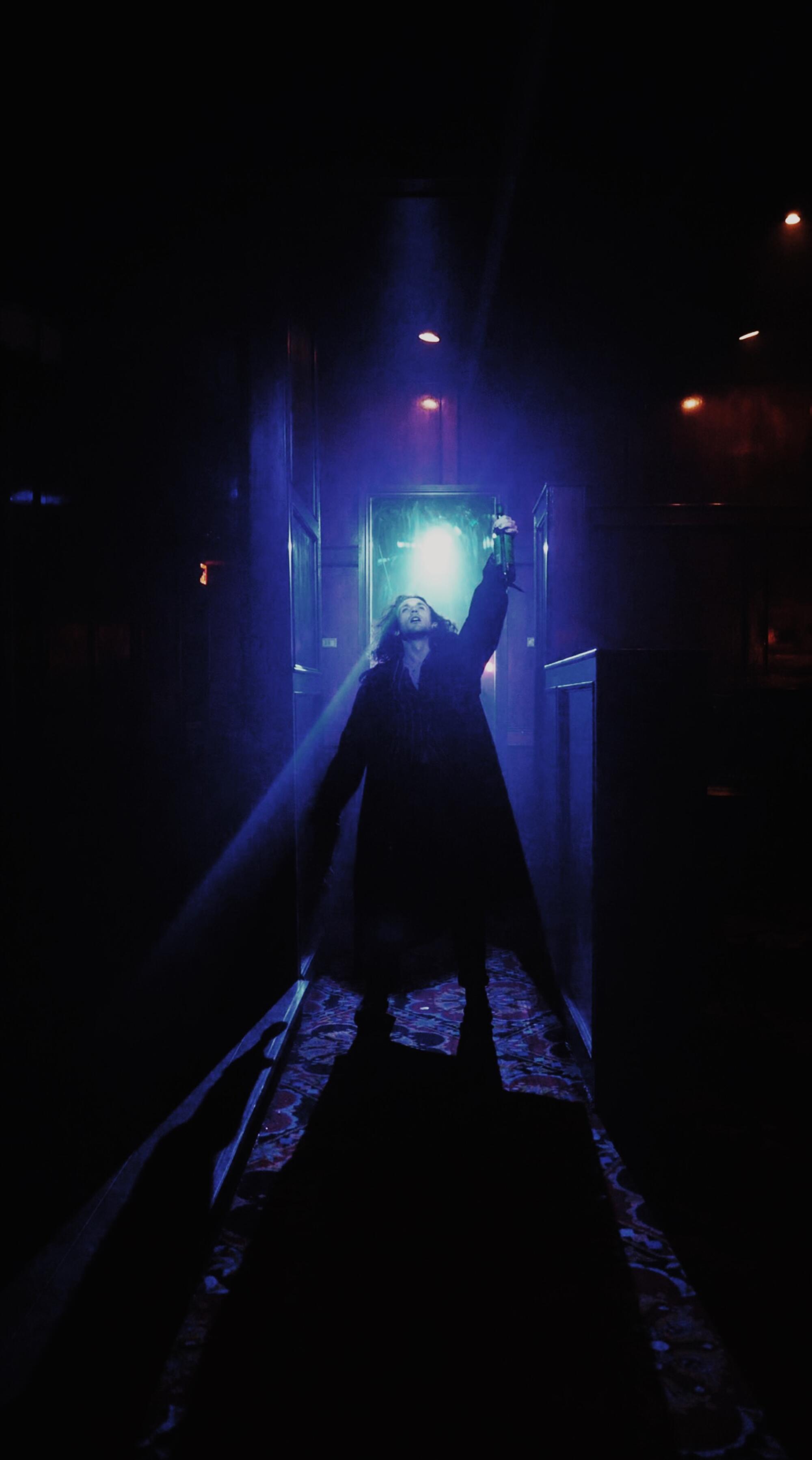 A figure in a long, dark coat stands in a dimly lighted hallway.