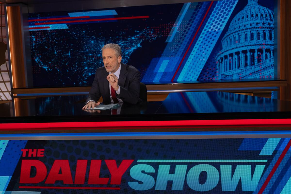 Jon Stewart sitting at a desk with 'The Daily Show' displayed across the front of it