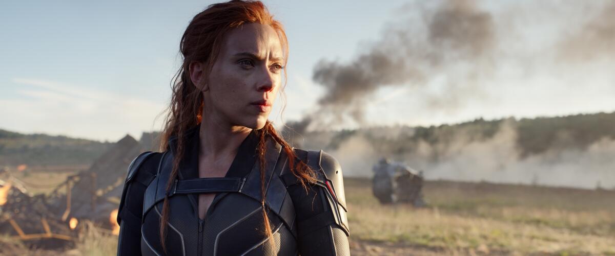 Scarlett Johansson in "Black Widow" looks stern before a background of smoldering vehicles.