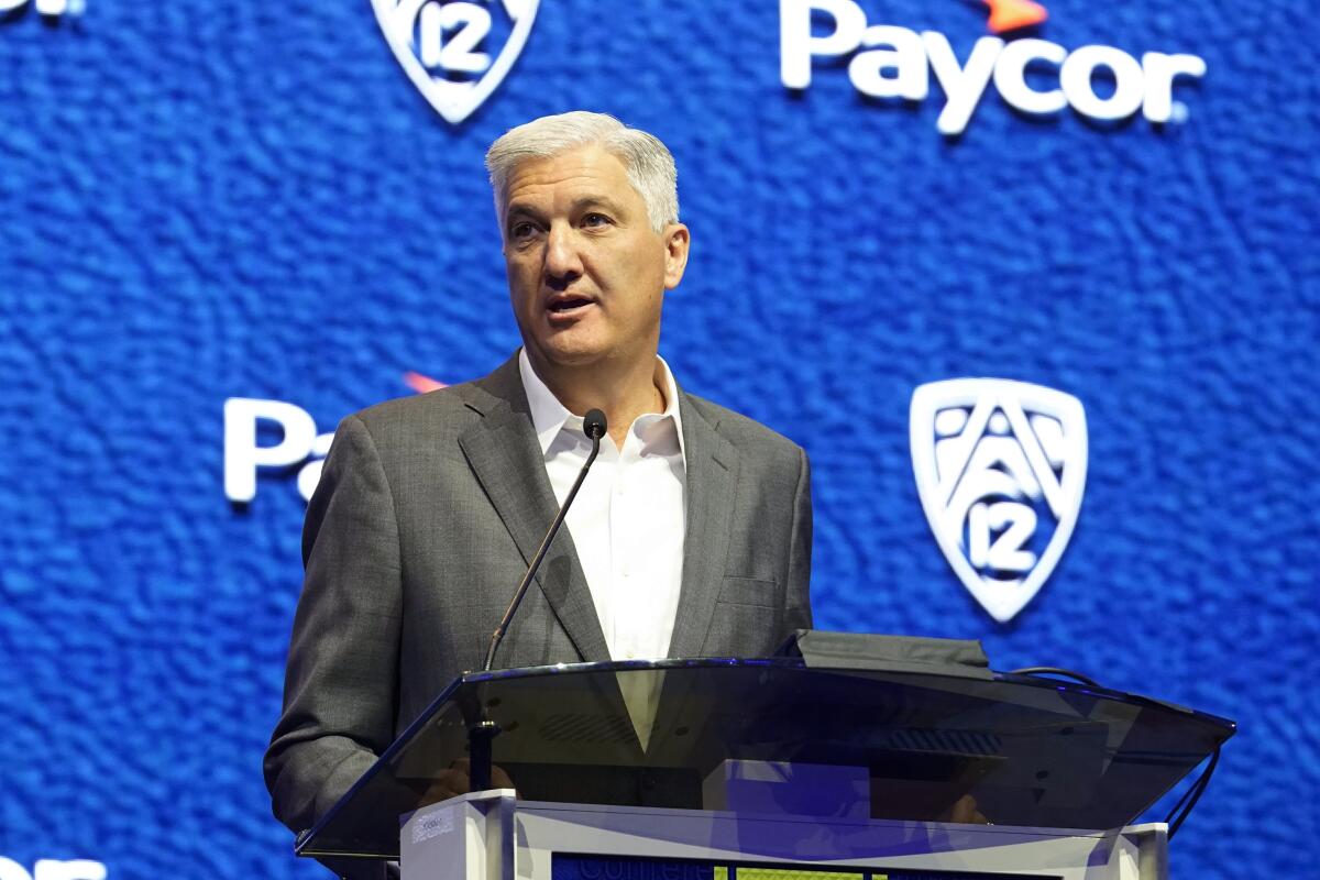 Pac-12 Commissioner George Kliavkoff speaks at a lectern in front of a bright blue background.