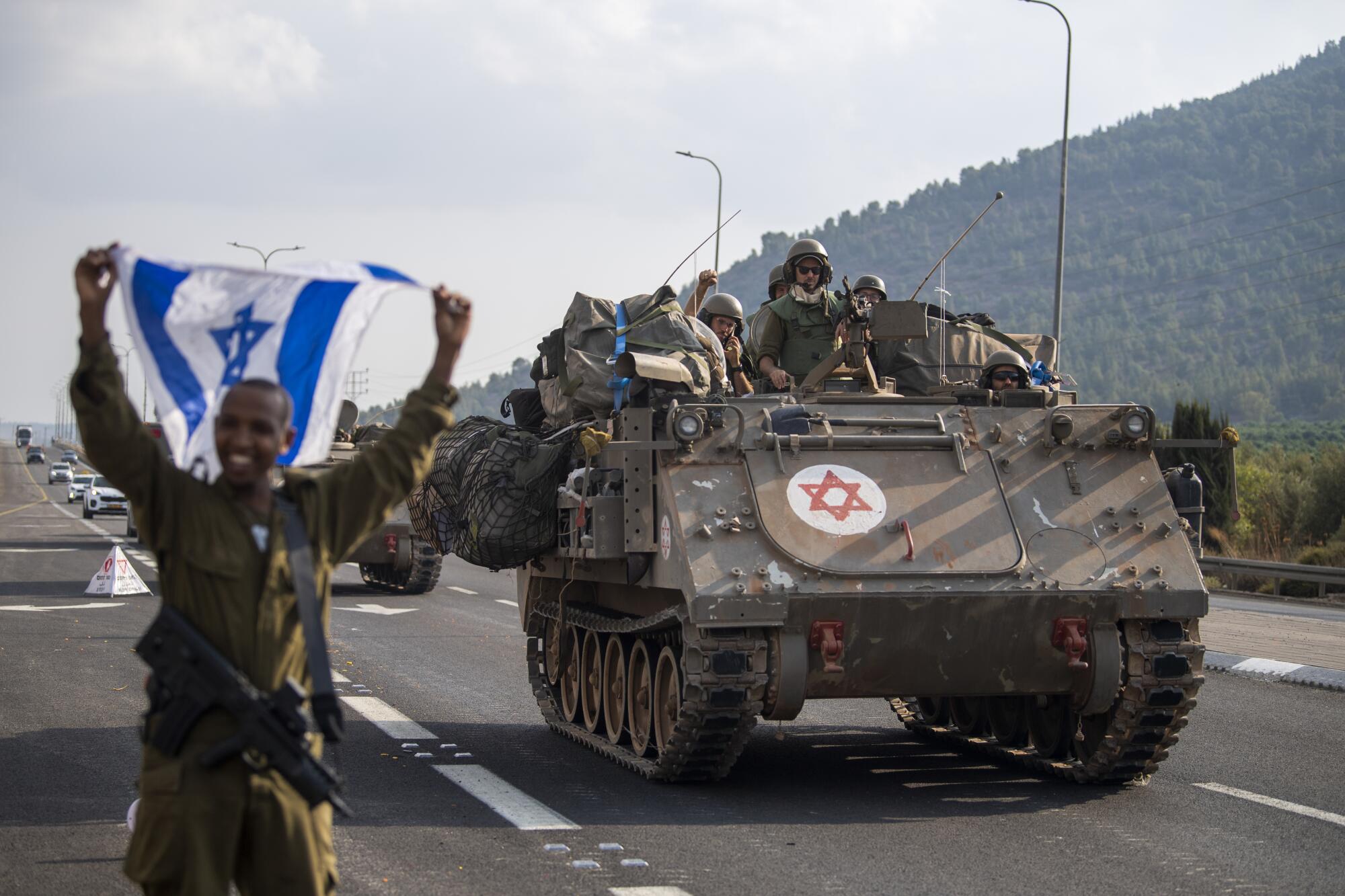 A man in fatigues holds up a blue-and-white flag as he stands on a road near an armored carrier with troops aboard 