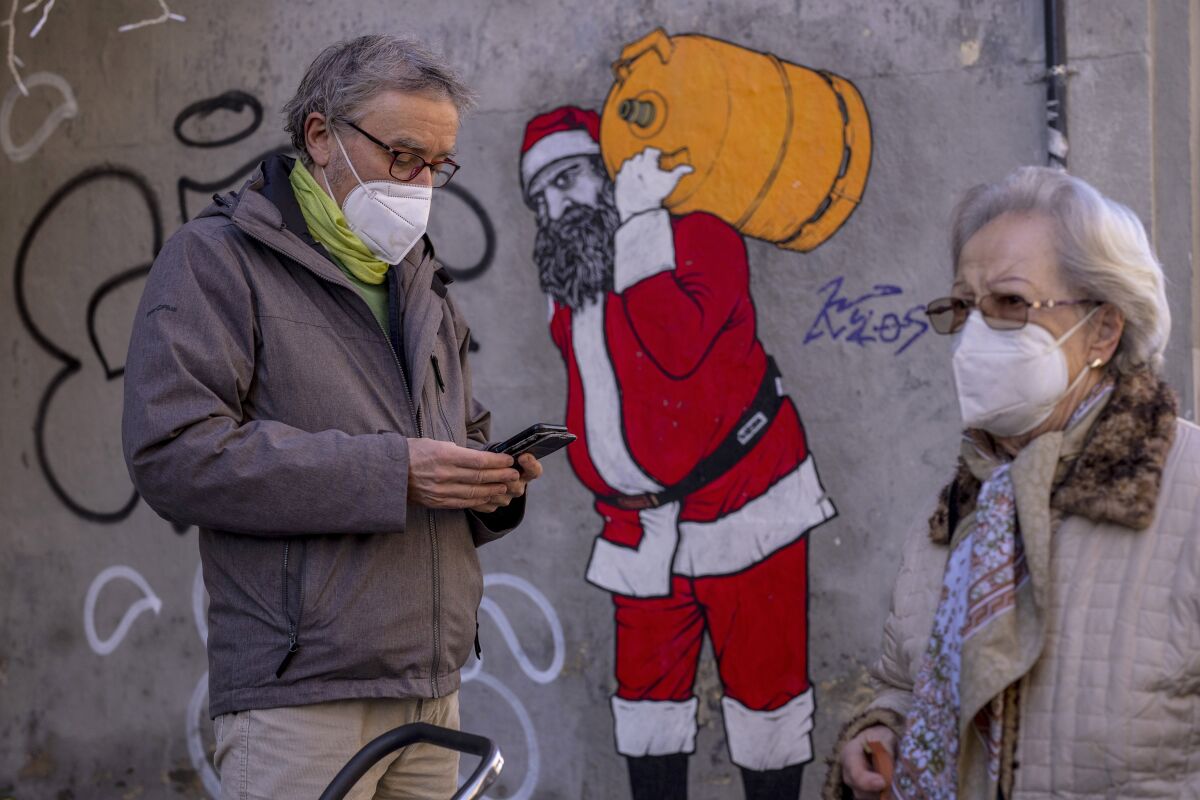 A man and woman wearing face masks walk on a street in front of a wall painted with a Santa Claus figure