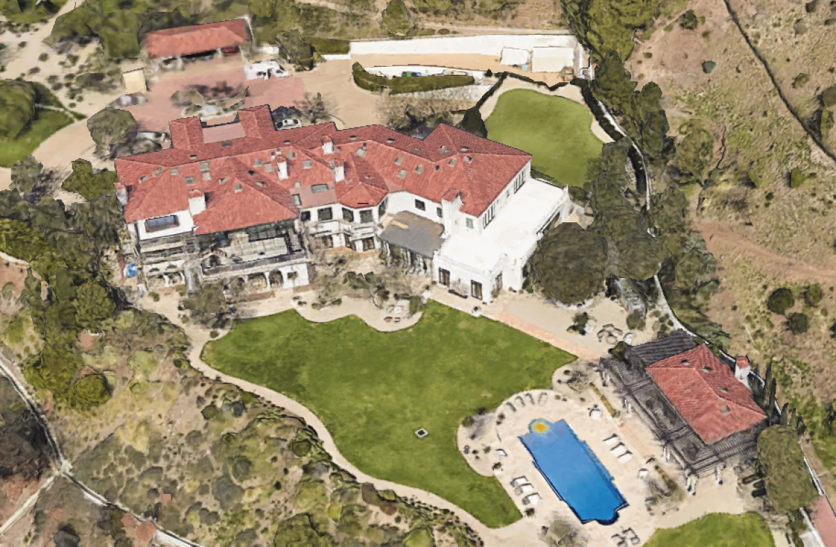 An aerial view of a property that includes a mansion, other buildings and a pool with lawn and trees surrounding the grounds.