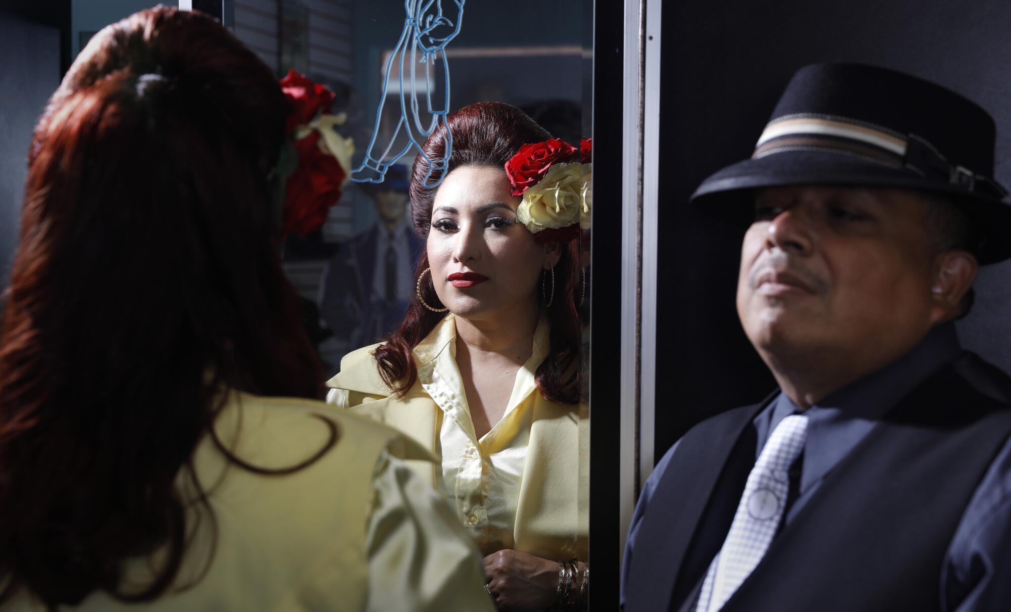 A woman wearing a 1940s hairdo and a bright yellow suit is seen in reflection next to a man wearing a dapper tie and hat