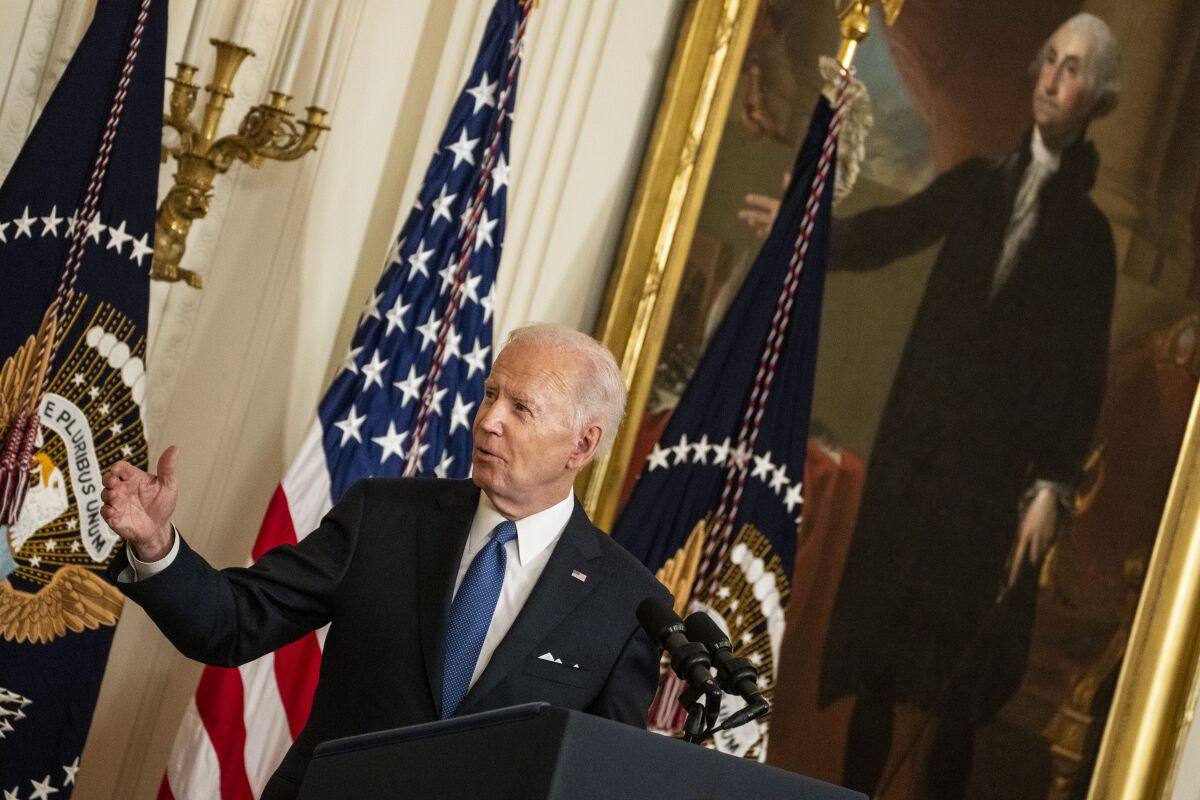 A man in a dark suit and blue tie gestures as he speaks against a backdrop of flags
