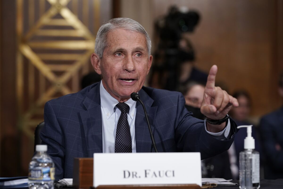 Dr. Anthony Fauci sits at a table with his name on a card and speaks into a microphone